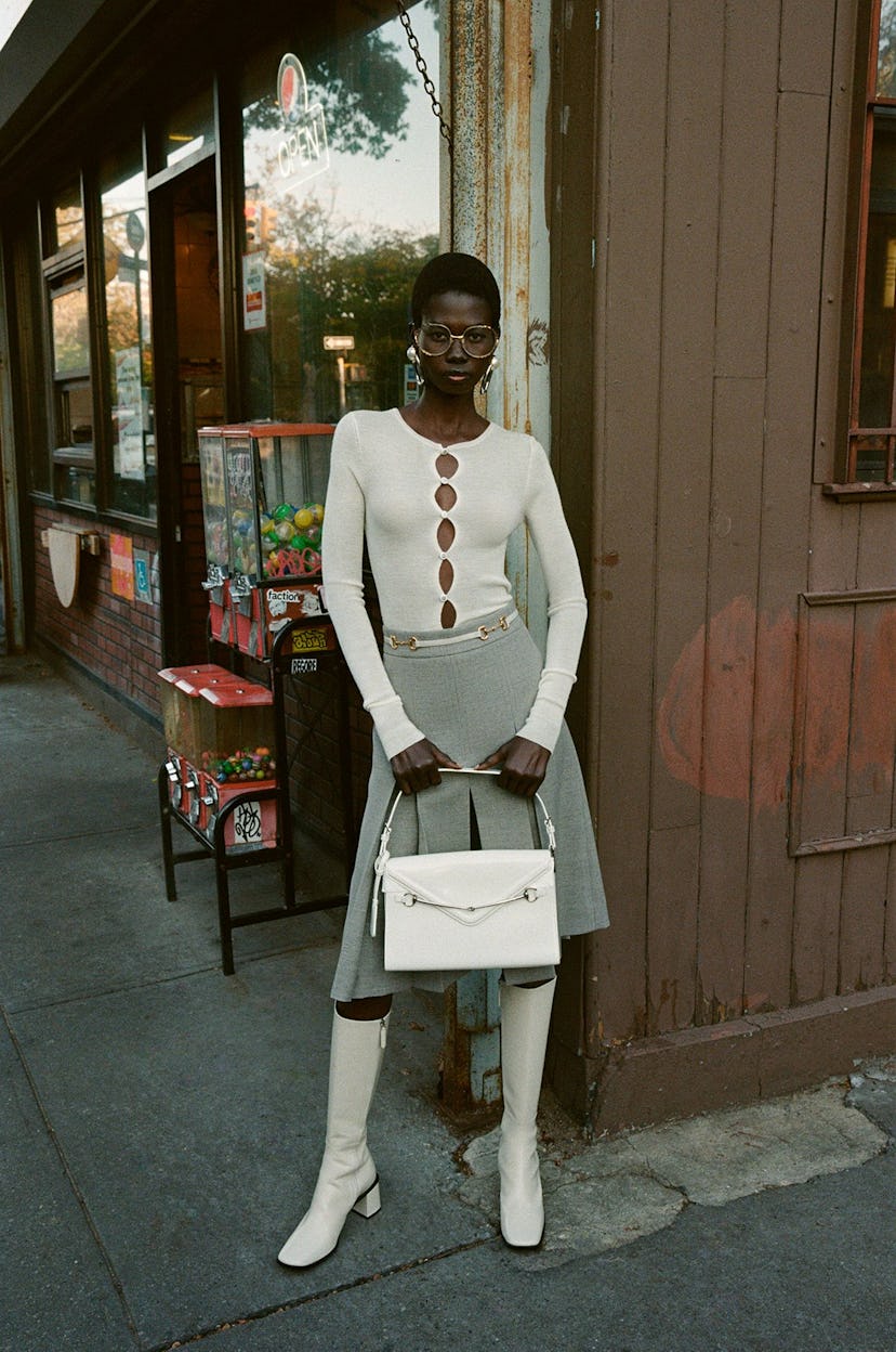 Stylish woman in a white cut-out top and grey skirt, paired with white boots and a bag, standing on ...